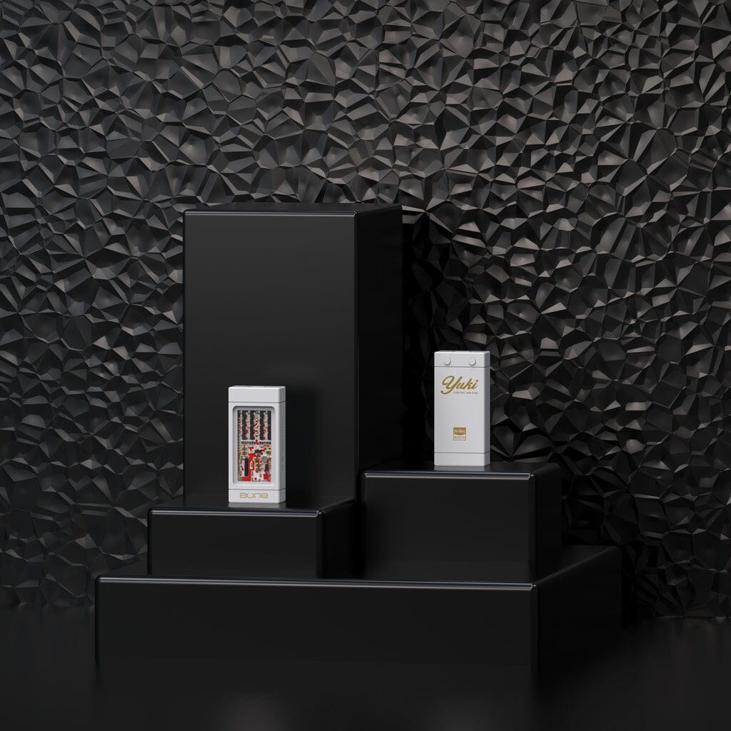 Two white product boxes are showcased on black geometric podiums against a textured black wall. One box features the gold Aune Audio YUKI branding, while the other displays vibrant graphics and text referencing the Aune Audio YUKI Portable DAC and Headphone Amplifier. The scene exudes a sleek, modern aesthetic.