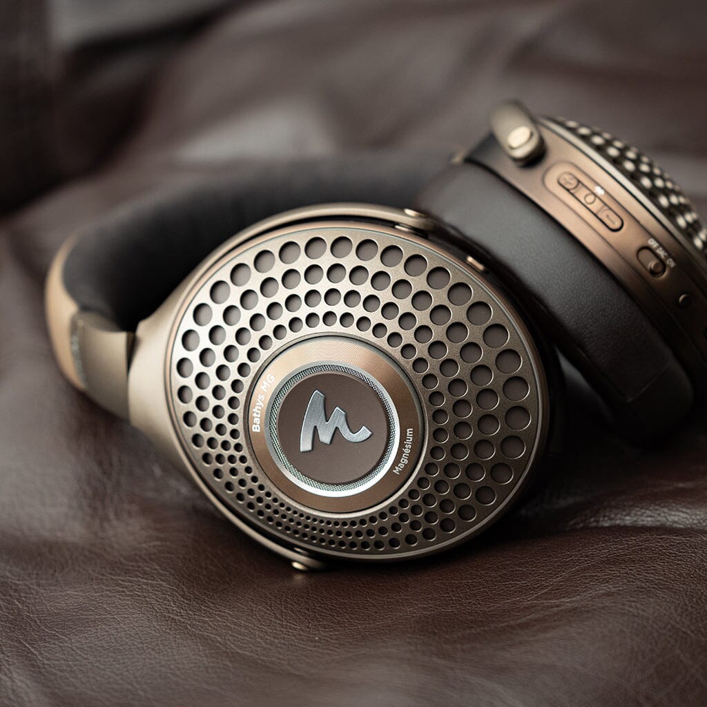 Close-up of Focal Bathys MG Wireless Headphones by Focal, featuring metallic circular perforated ear cups, visible buttons, and a cushioned headband, resting on a brown leather surface.