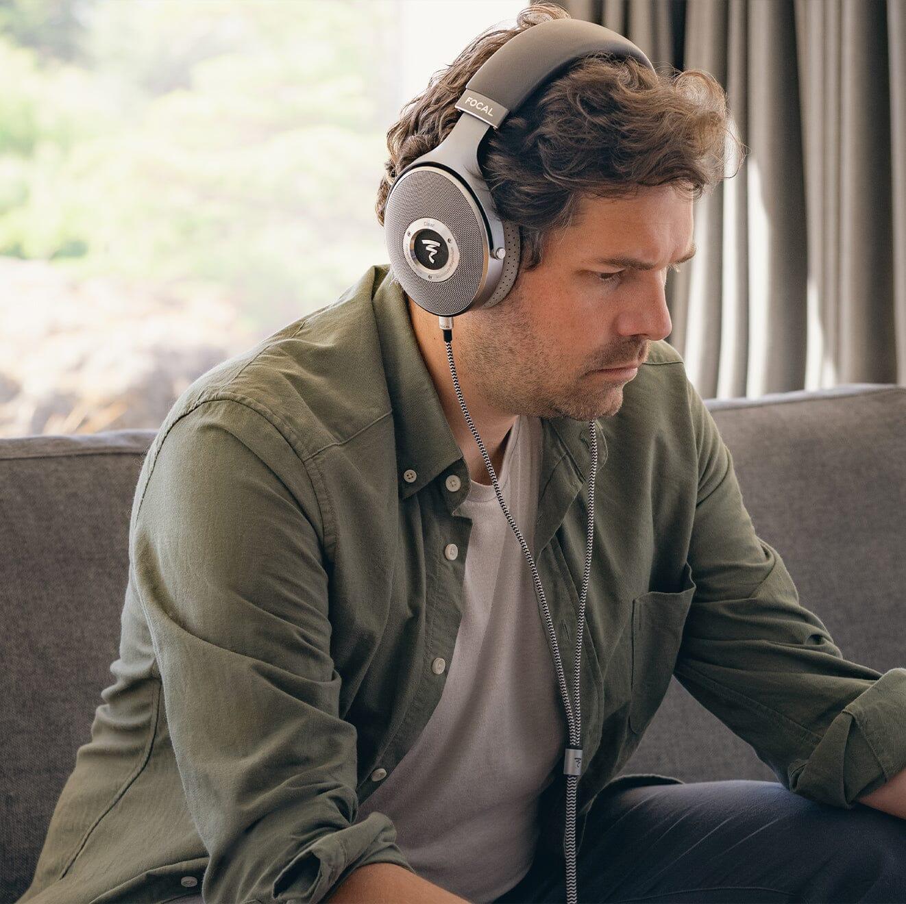A man in a green button-up over a white t-shirt sits on a couch, focused, wearing Focal Clear Headphones by Focal from headphones.com. Greenery is visible through the window behind him.