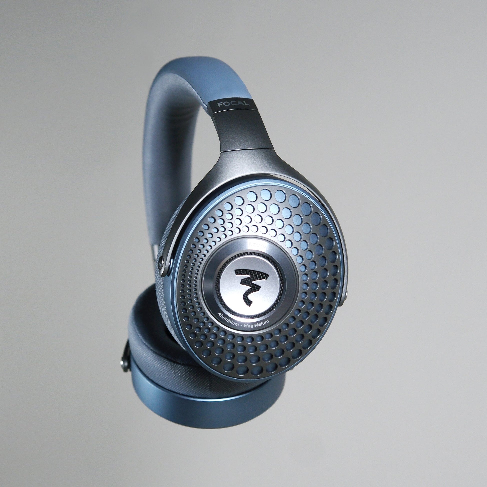 The Focal Azurys by Focal are closed-back headphones in metallic blue, featuring a patterned grille and bold circular logo on the ear cup, shown suspended against a plain light gray background.
