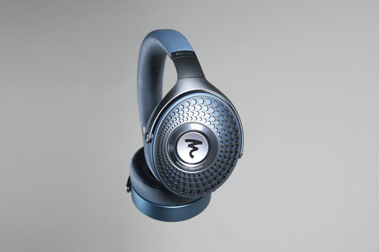 The Focal Azurys by Focal are closed-back headphones in metallic blue, featuring a patterned grille and bold circular logo on the ear cup, shown suspended against a plain light gray background.