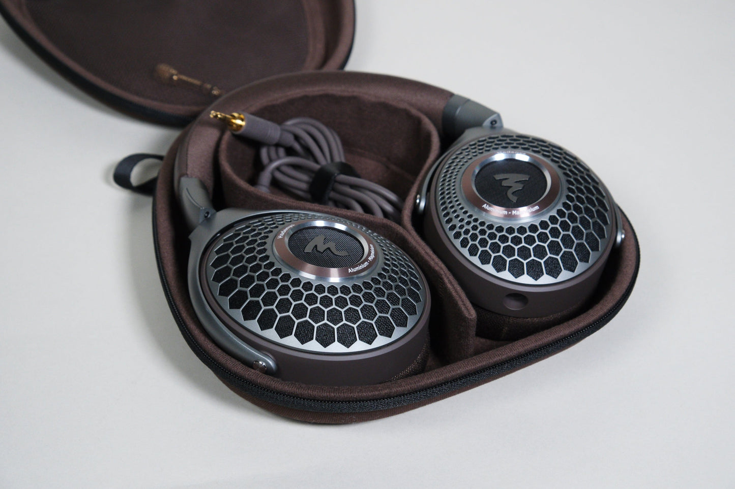 The Focal Hadenys headphones by Focal, featuring open-back honeycomb-patterned ear cups and a coiled cable, are neatly stored in a brown protective carrying case.
