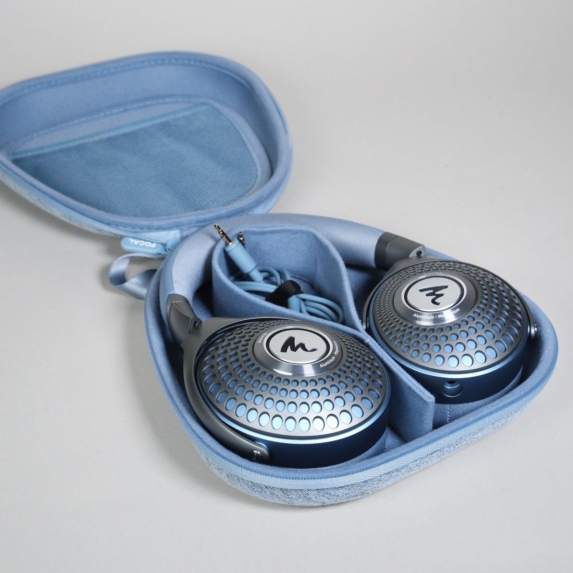A pair of metallic blue Focal Azurys closed-back headphones with a geometric pattern rests in a light blue zippered carrying case, with the headphone cable neatly coiled inside.
