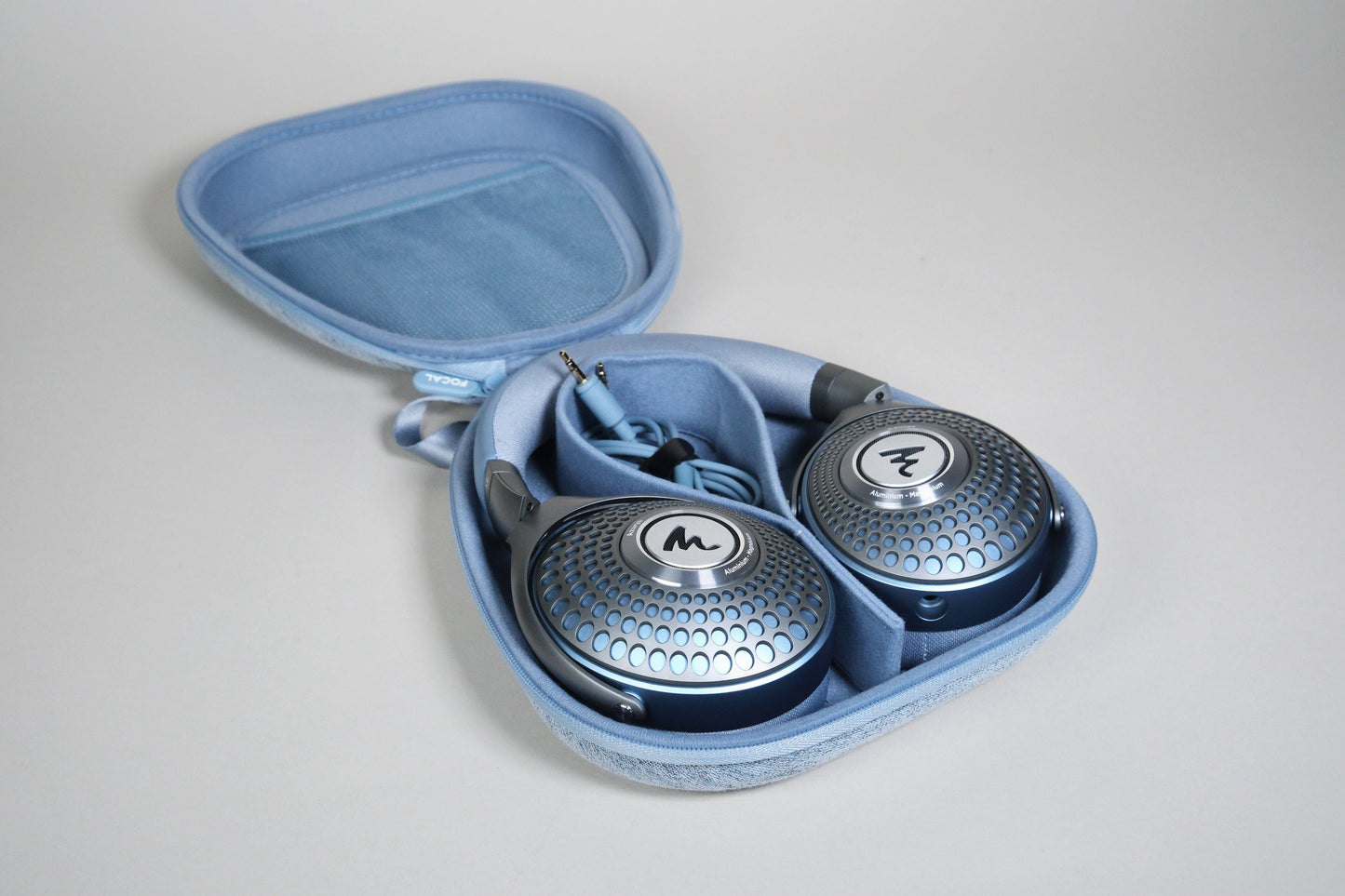 A pair of metallic blue Focal Azurys closed-back headphones with a geometric pattern rests in a light blue zippered carrying case, with the headphone cable neatly coiled inside.