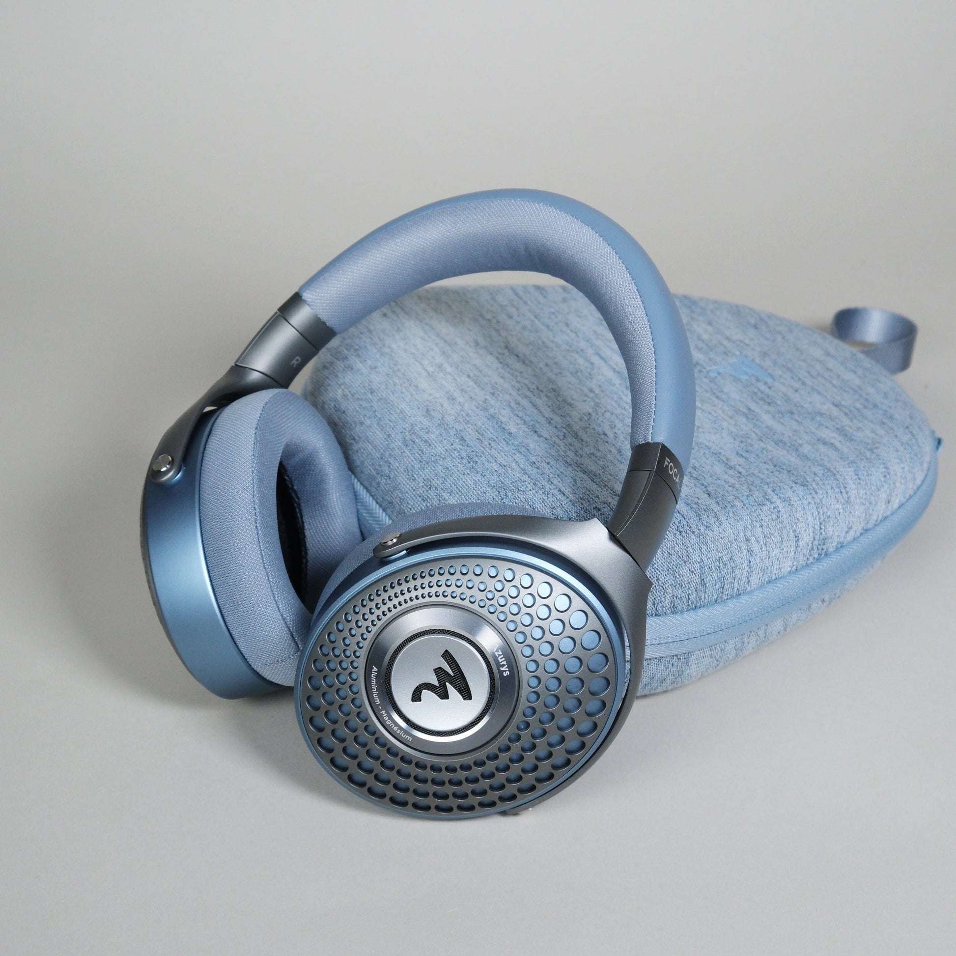 A pair of Focal Azurys closed-back headphones by Focal, featuring blue ear cups with a circular textured design, rest on a matching blue fabric case against a light gray background.