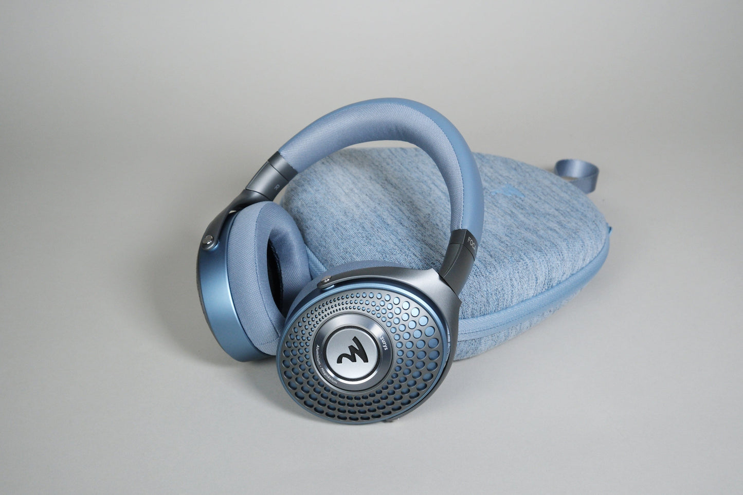 A pair of Focal Azurys closed-back headphones by Focal, featuring blue ear cups with a circular textured design, rest on a matching blue fabric case against a light gray background.