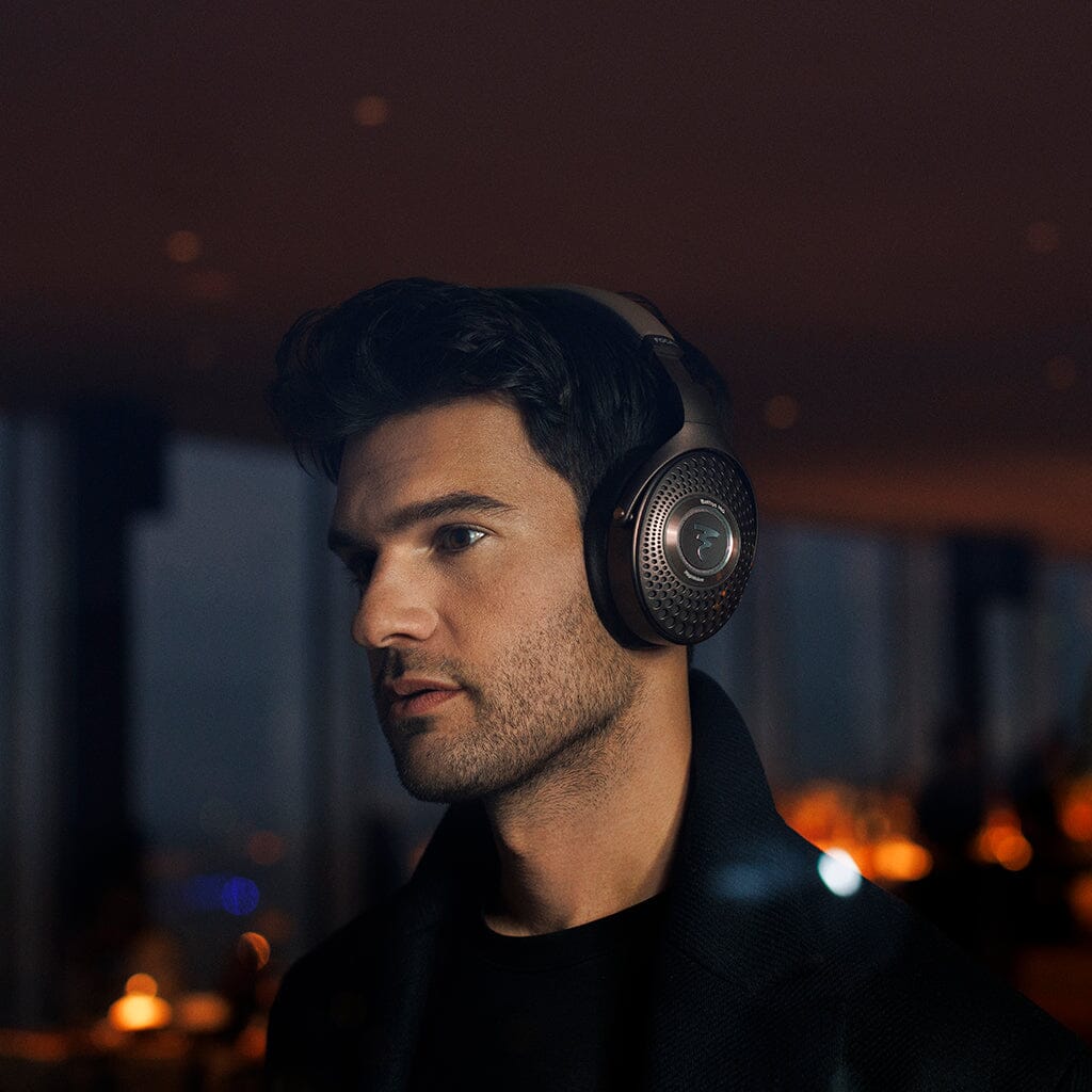 A man with dark hair and light stubble wears the Focal Bathys MG Wireless Headphones by Focal indoors, set against a softly lit, blurred background that evokes a modern, upscale atmosphere.