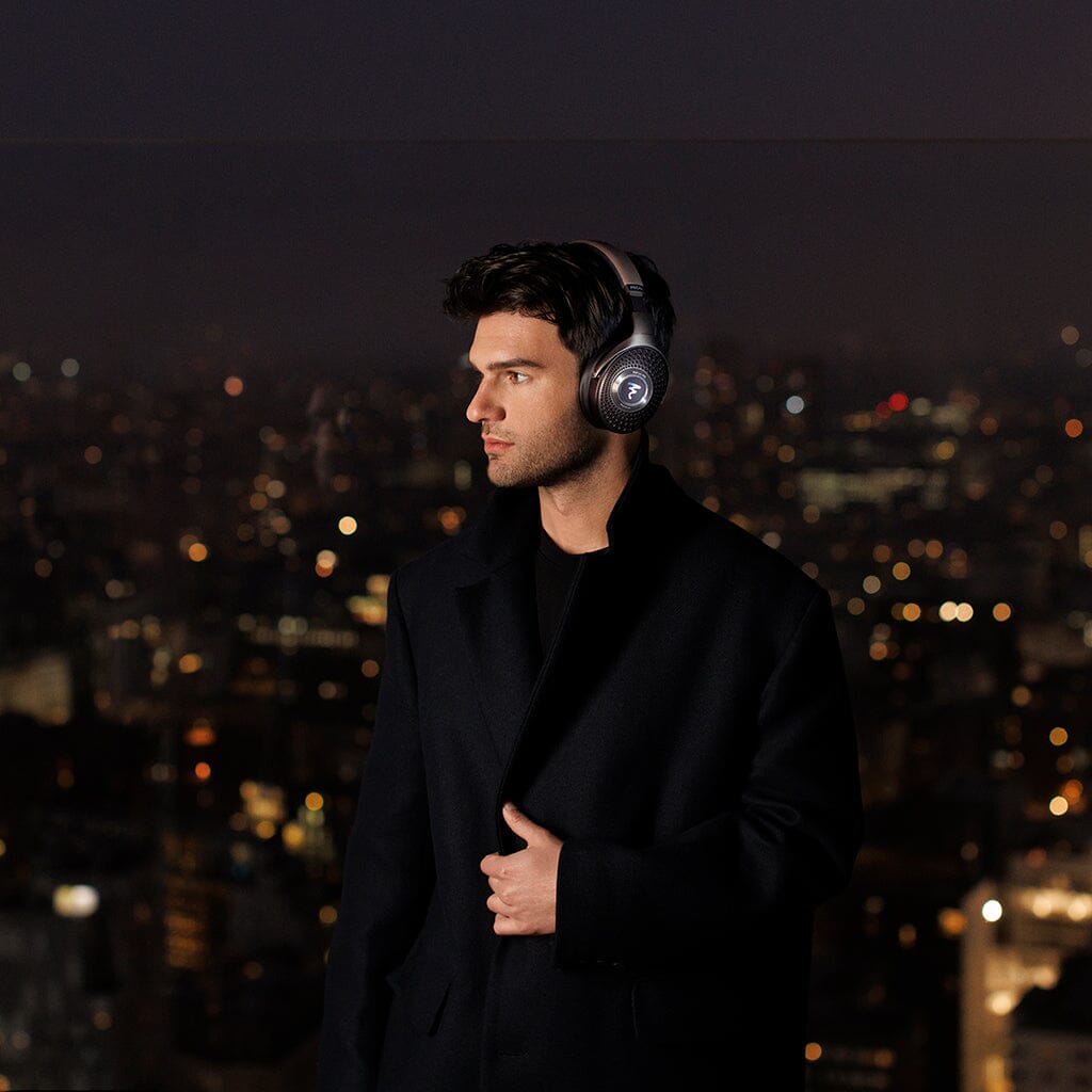 A man wearing Focal Bathys MG Wireless Headphones and a dark coat stands against a blurred cityscape at night, illuminated by city lights in the background.