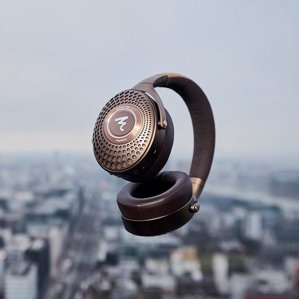 A pair of Focal Bathys MG Wireless Headphones by Focal, featuring a metallic finish, floats in mid-air with a blurred cityscape in the background.