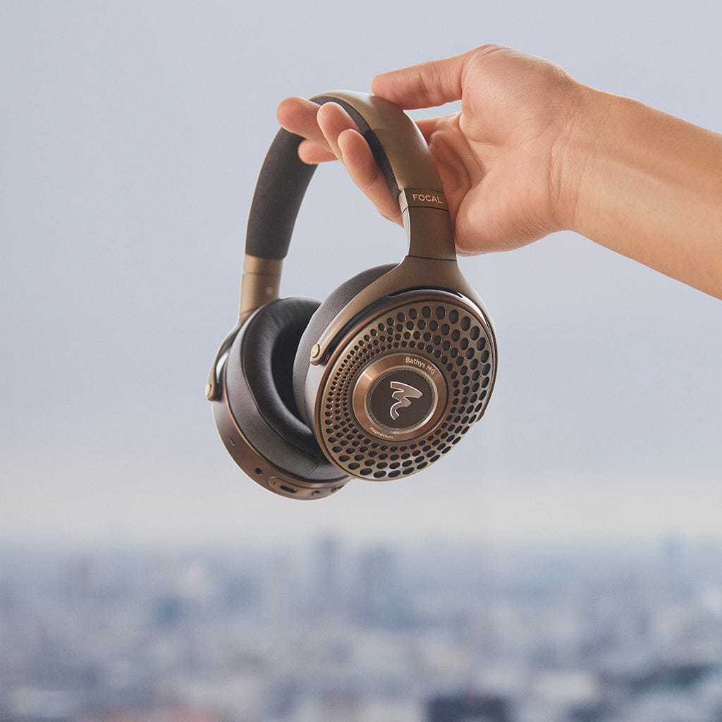 A hand holds up a pair of Focal Bathys MG Wireless Headphones by Focal, featuring a circular design, against a blurred cityscape background.