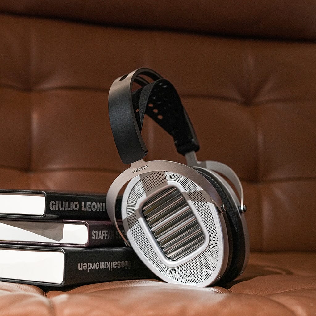 A pair of large, silver and black Hifiman Ananda Unveiled Headphones sits on a stack of books on a brown leather couch, highlighting the modern Hifiman design with distinct open-back metal grilles.