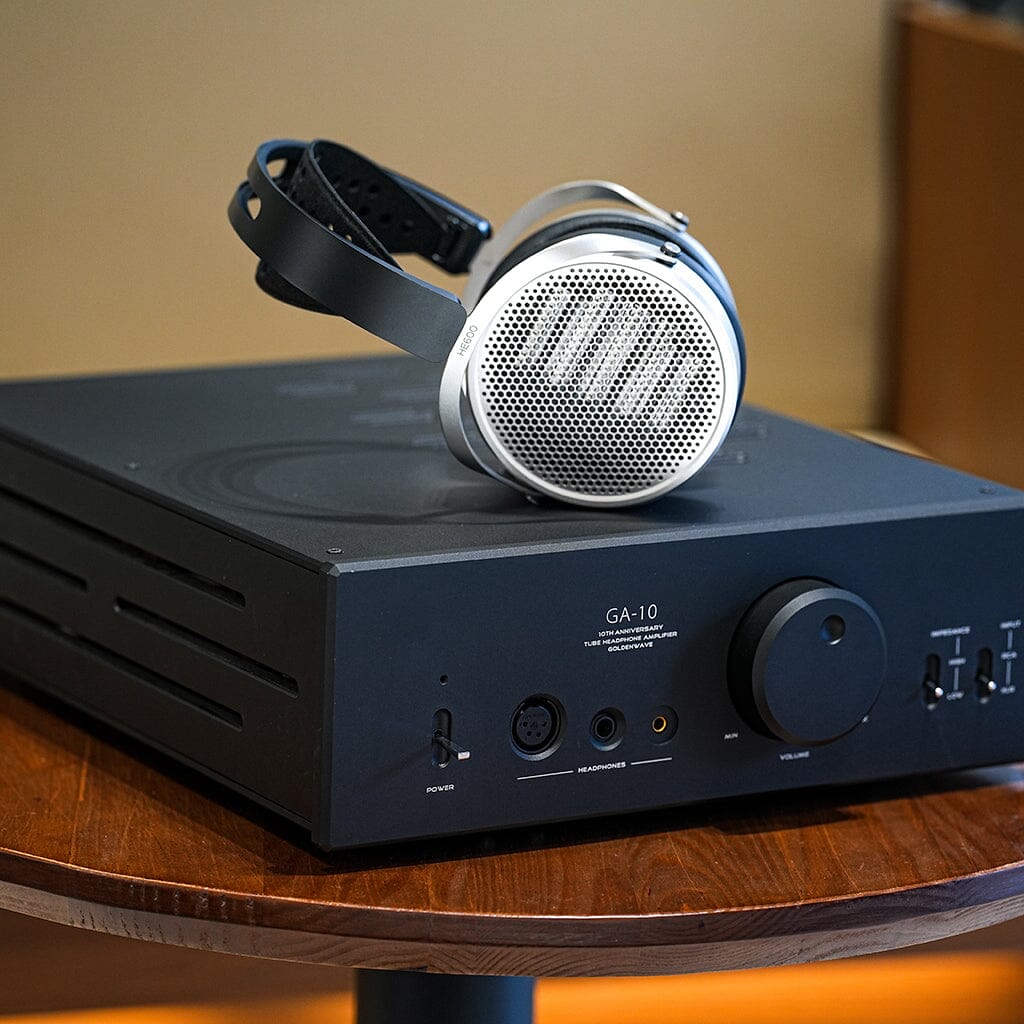 A pair of silver Hifiman HE600 headphones from Hifiman sits atop a black GA-10 headphone amplifier with knobs, buttons, and input ports on its front panel, all arranged on a wooden table.