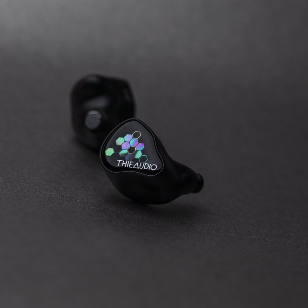 A pair of Thieaudio Monarch MKIV In-Ear Headphones with glossy, logo-embellished faceplates and geometric multicolored designs lies on a dark gray surface.