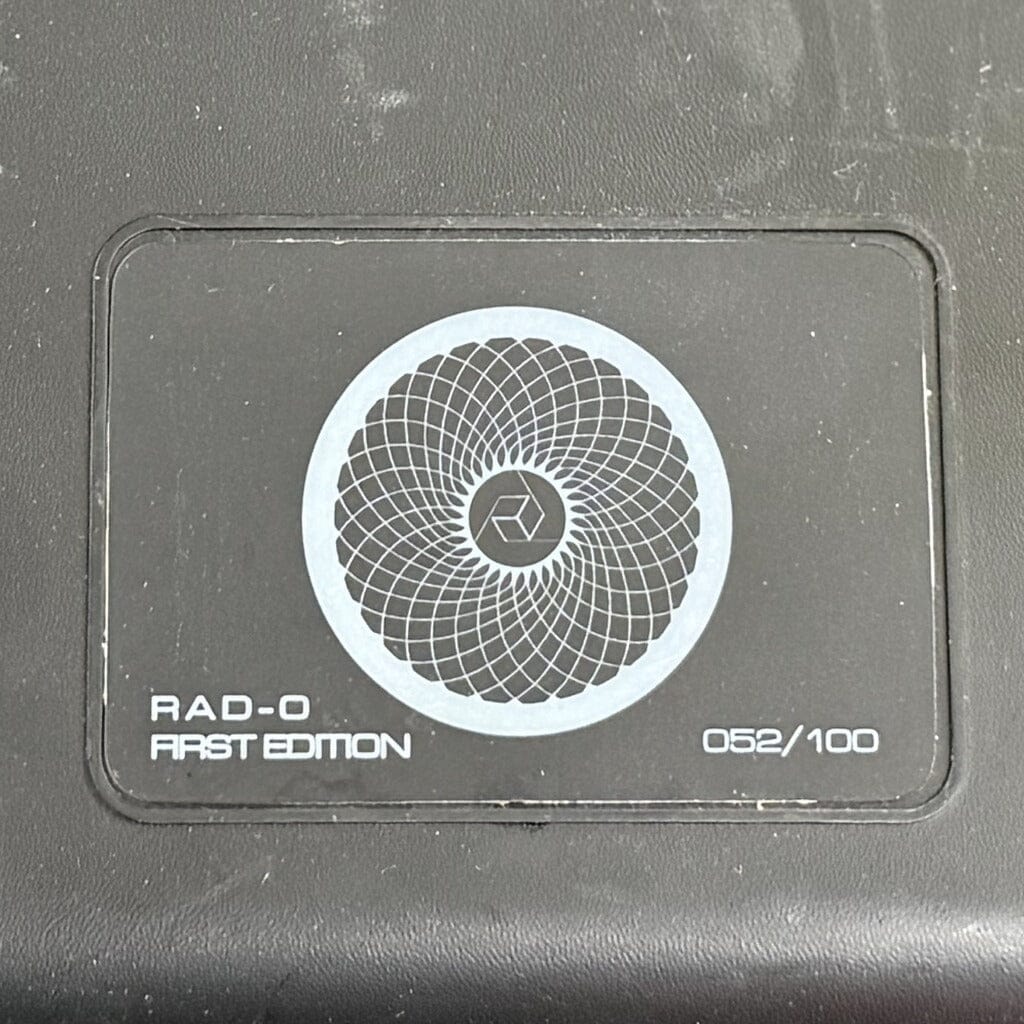 A sleek black rectangular label features a central geometric circular pattern, the text "RAD-0 FIRST EDITION," and the serial number 052/100 on the bottom right. The dark, textured background captures the refined aesthetic of Rosson Audio Design’s RAD-0 Headphones—Open Box Clearance unit 52.