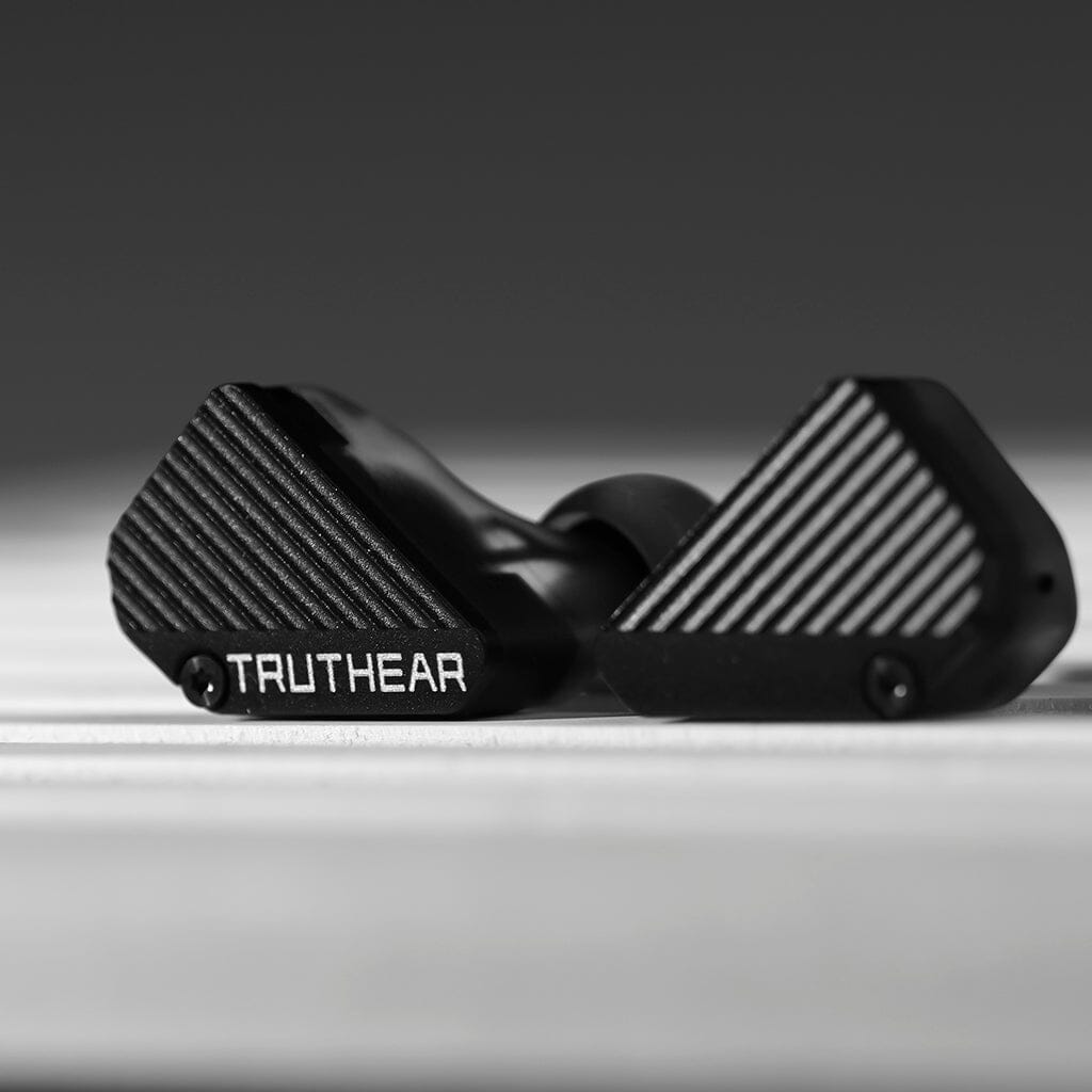 Close-up of two black, triangular Truthear Pure In-Ear Headphones by TRUTHEAR, featuring ridged surfaces and brand name, resting on a white slatted surface with a dark background—highlighting exceptional sound quality.