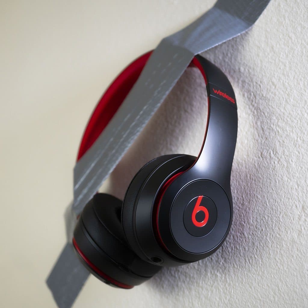 A pair of black and red Mediocre Headphones on Wall by headphones.com is taped to a beige wall with duct tape—an unconventional display.