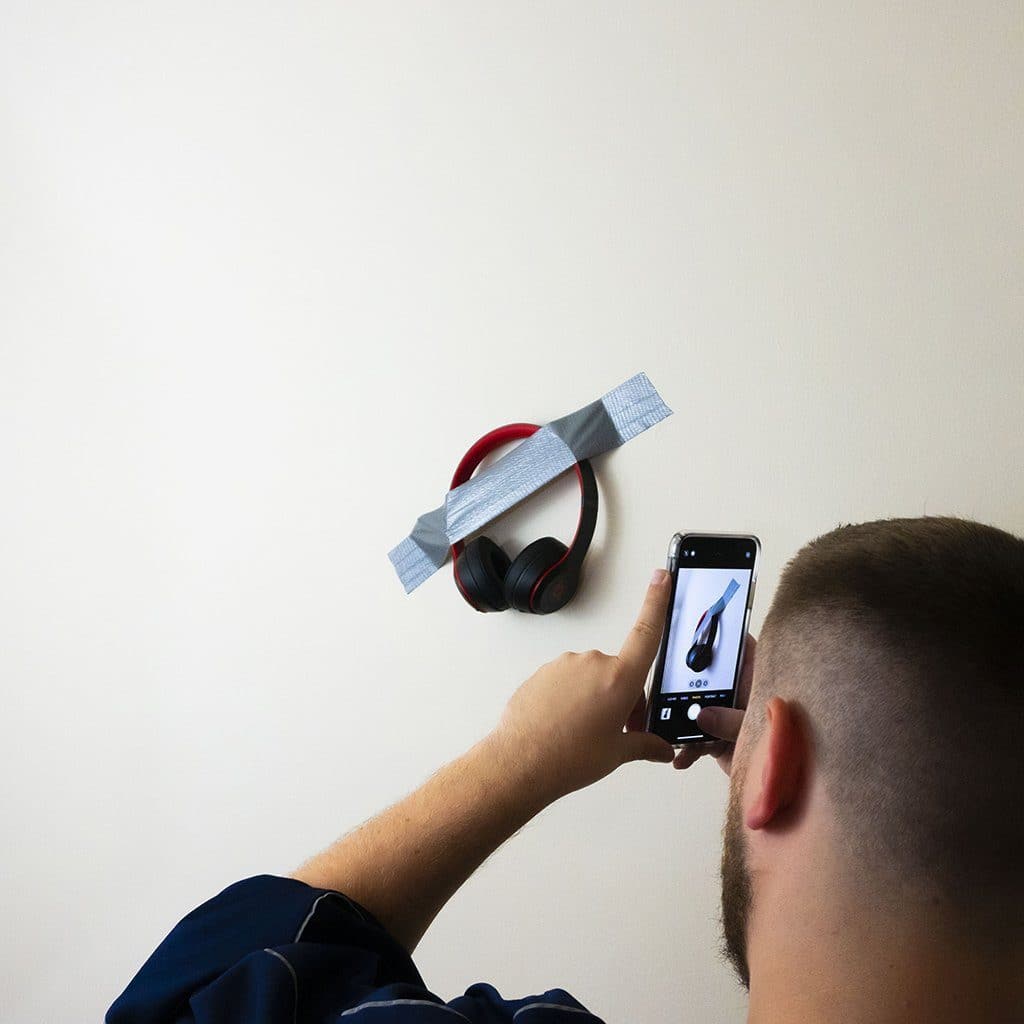 Someone snaps a photo with their smartphone of Mediocre Headphones on Wall by headphones.com, taped to a white wall like modern art—a quirky tribute from headphones.com.