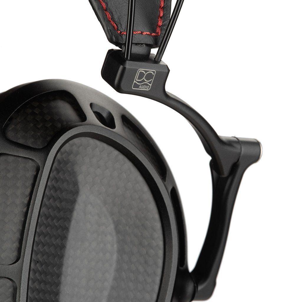 Close-up of a black ear cup with carbon fiber texture, metal and leather accents, red stitching, and Dan Clark Audio Stealth logo—highlighting the planar magnetic Dan Clark Audio Stealth Headphones - Open Box.