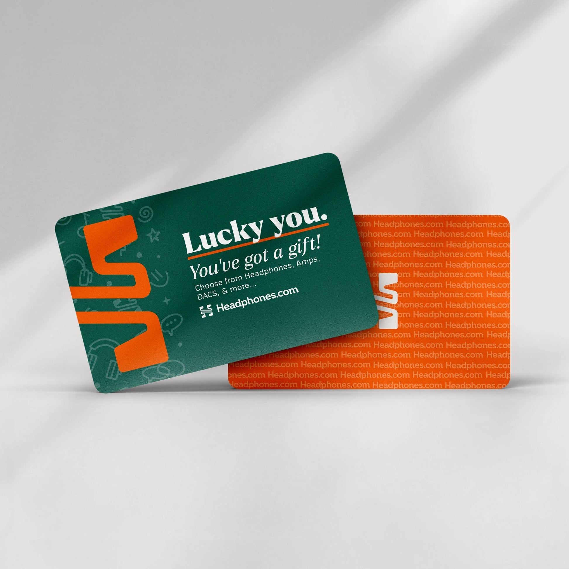 Two headphones.com Gift Cards are shown—one reads, “Lucky you. You’ve got a gift! Choose from headphones, amps, DACs & more.” Redeem easily and enjoy the repeating orange headphones.com pattern on the second card.