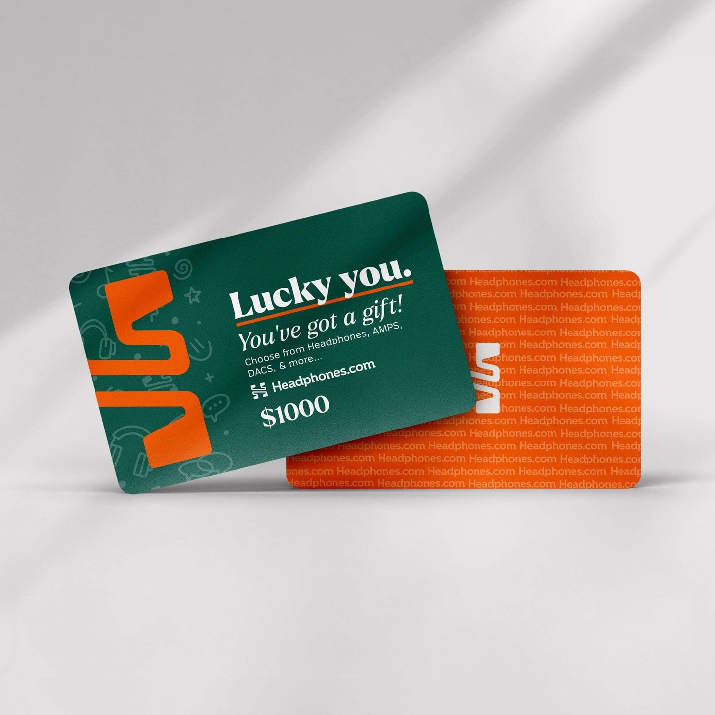 A green headphones.com gift card with “Lucky you. You’ve got a gift!” in white and $1000 displayed is next to an orange Headphones.com card, both resting on a white surface—ready to redeem for your next audio upgrade.