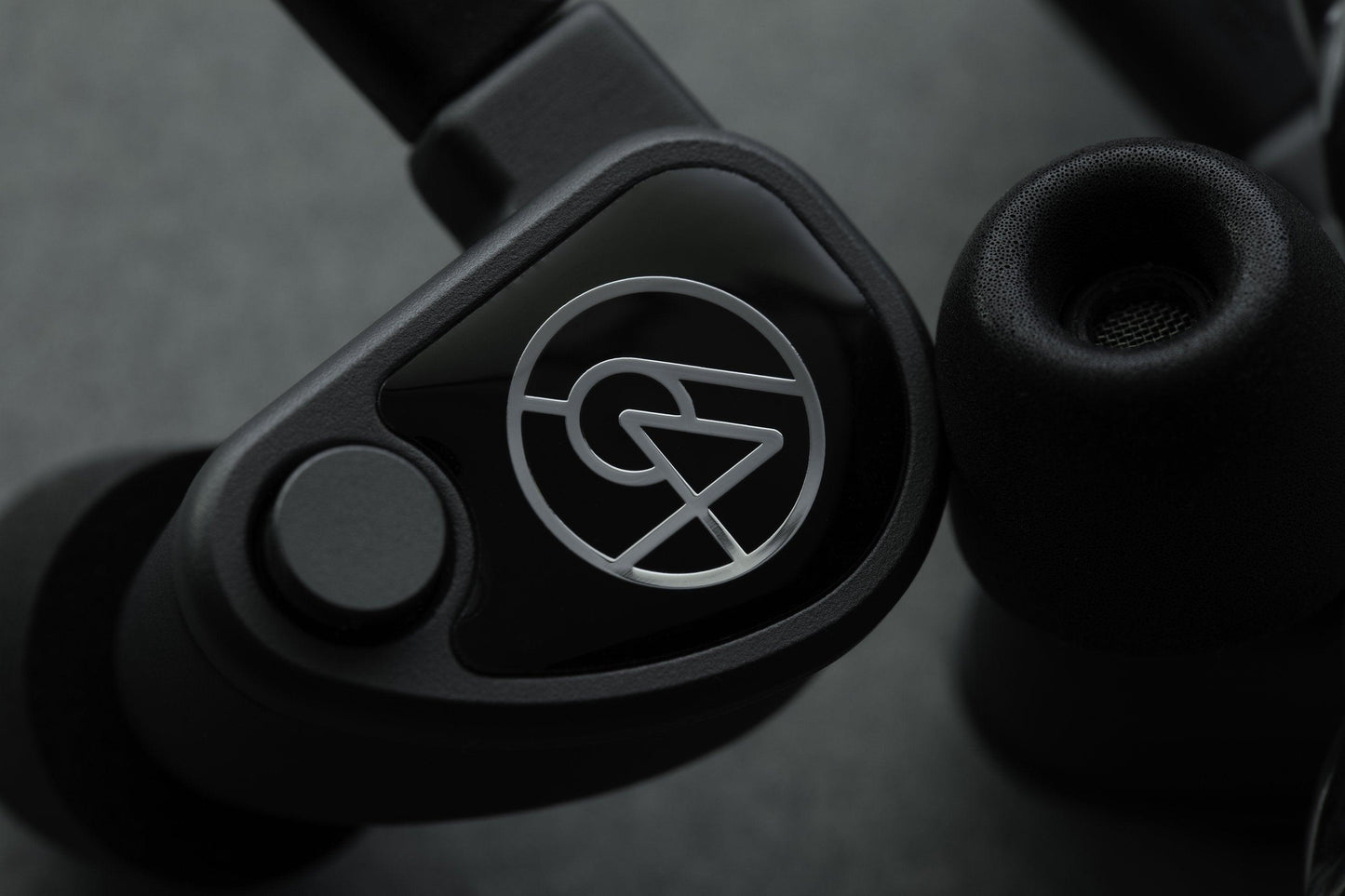 Close-up of the 64 Audio U6t In-Ear Headphones - Open Box, featuring a black ergonomic aluminum shell with an abstract circular silver logo, by 64 Audio, resting on a grey textured background.