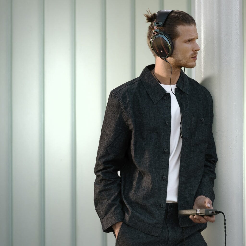 A man with long hair tied back wears large Meze Strada headphones and a dark jacket over a white shirt, holding a portable device against a light metal wall, enjoying impressive tonal balance.