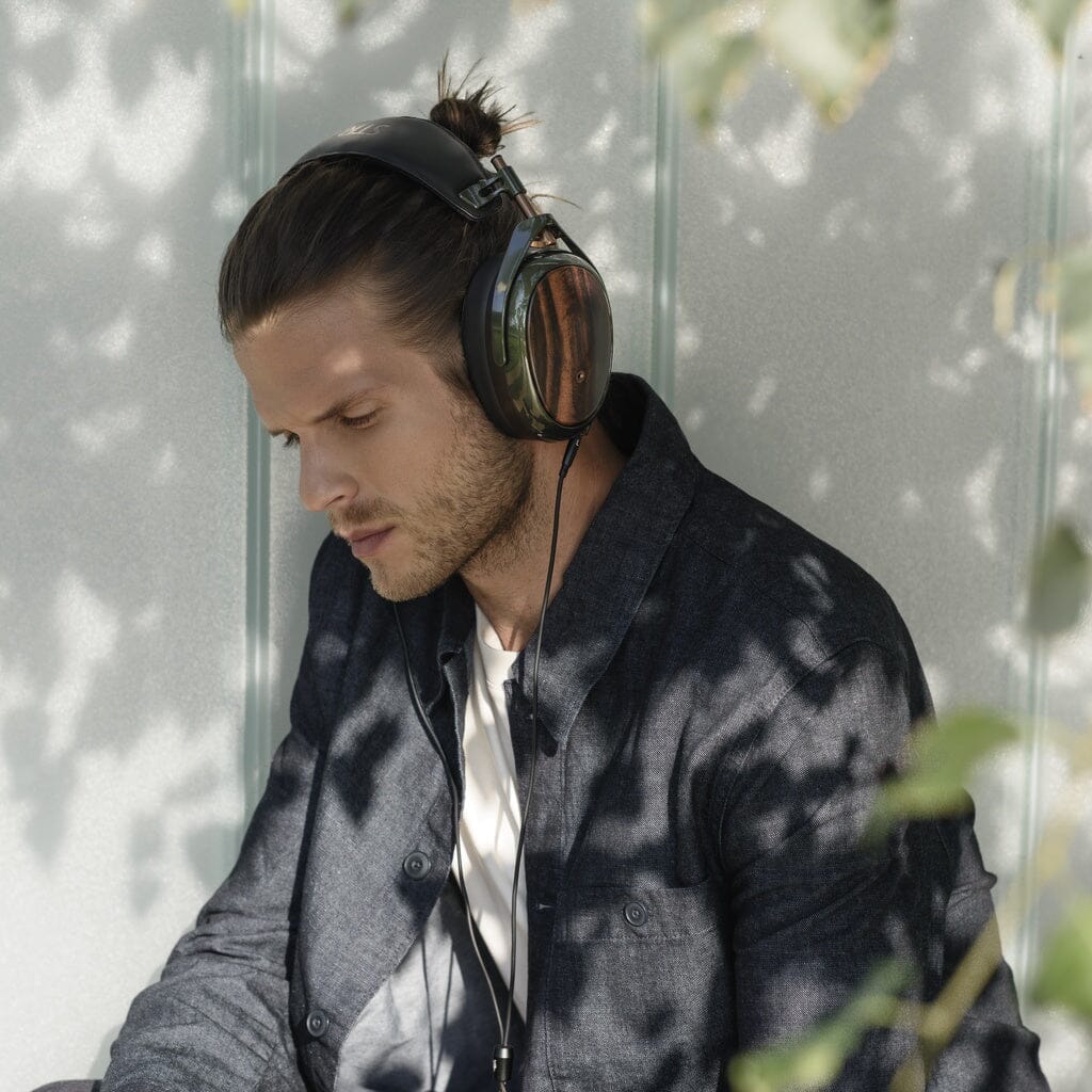 A man with long hair tied back wears Meze Strada closed-back headphones and a dark jacket, sitting outdoors in dappled sunlight among leafy shadows—enjoying perfect tonal balance.