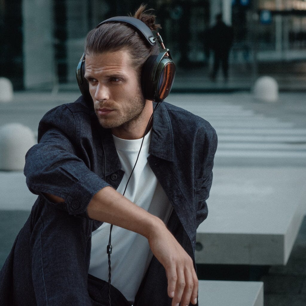 A man with long hair tied back wears Meze Strada Headphones and a dark jacket over a white shirt, sitting outdoors on a modern concrete surface, looking thoughtfully to the side as he enjoys the headphones' perfect tonal balance.