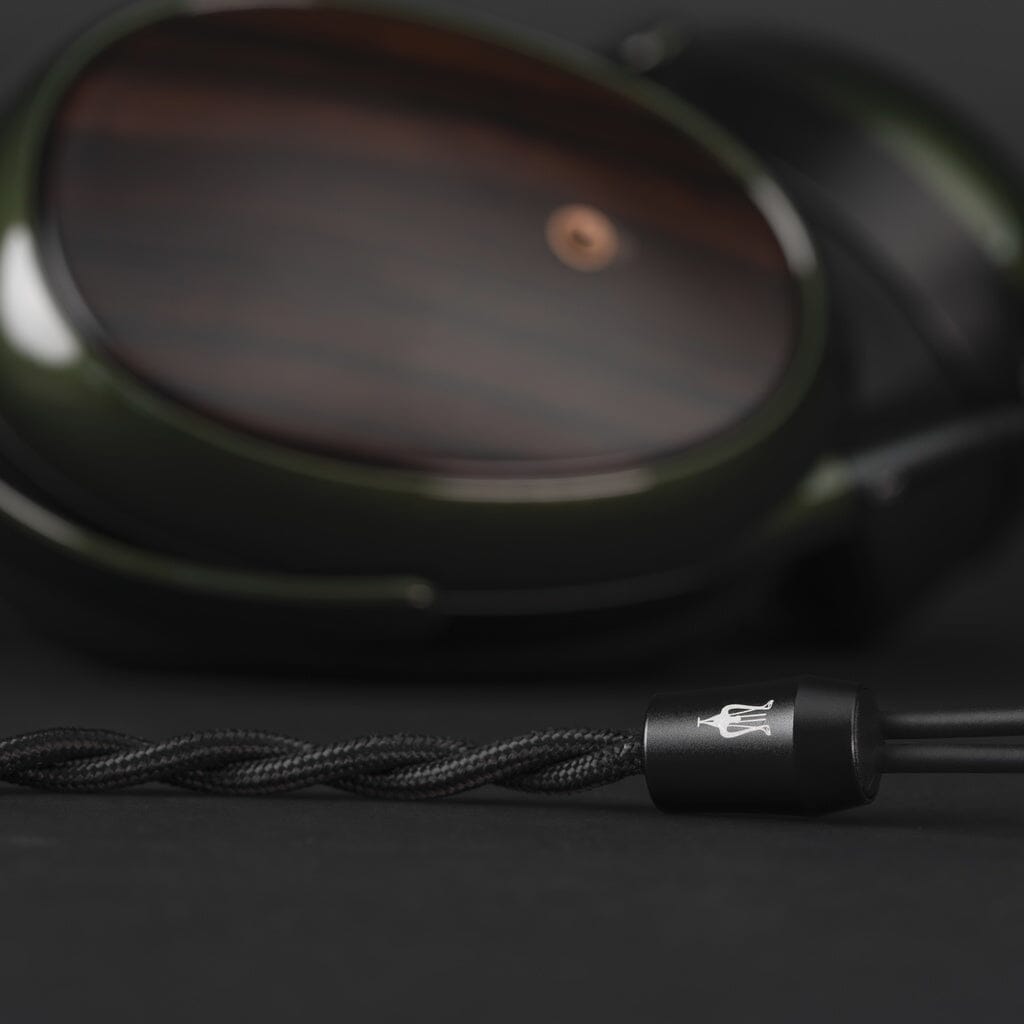 Close-up of Meze Audio Strada closed-back dynamic headphones with dark wood ear cups and a braided black cable on a dark background. A small, silver Meze logo is visible on the connector, reflecting the brand's tonal balance commitment.