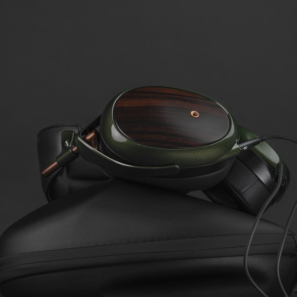 A pair of Meze Audio Strada closed-back headphones with dark green and wood-finish ear cups rests atop a black carrying case, showcasing Meze’s superb tonal balance against a dark background.