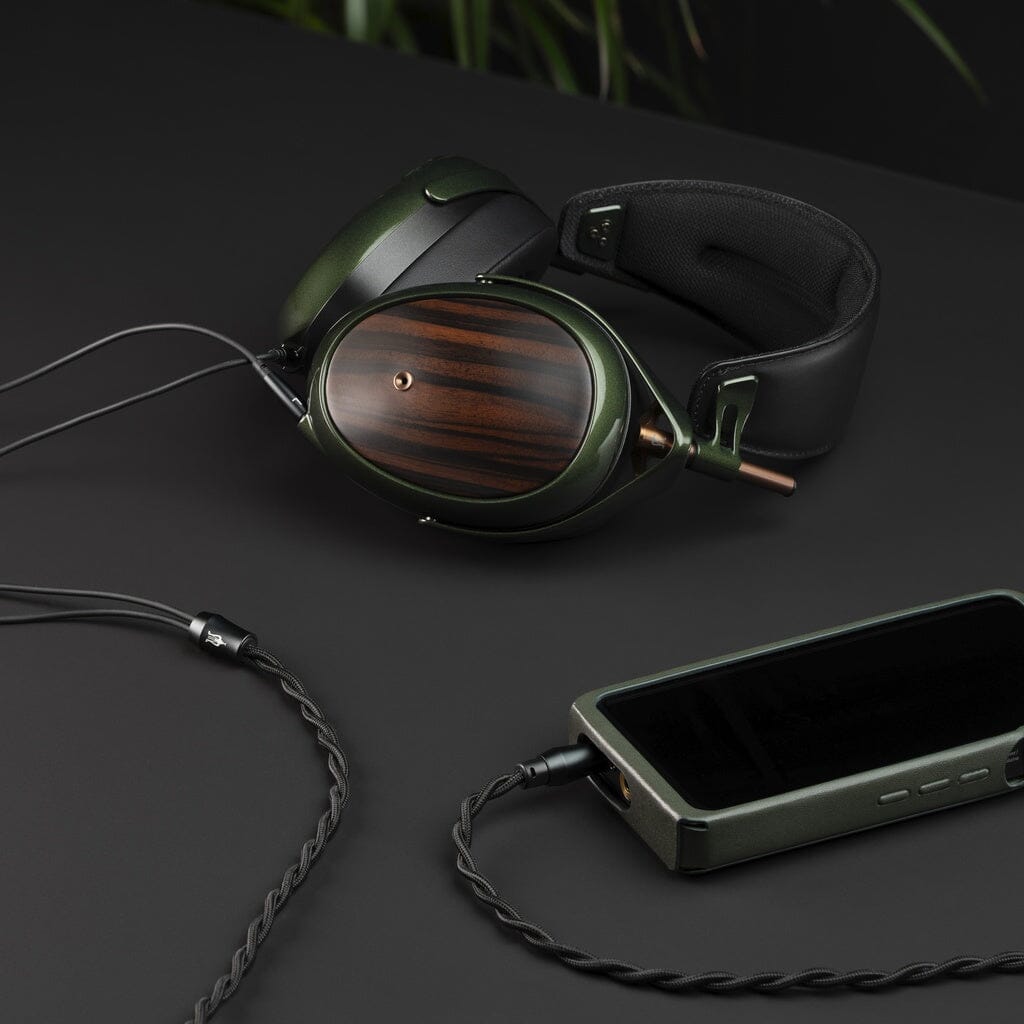 Meze Audio Strada closed-back dynamic headphones in dark green and wood grain with a black padded headband are connected by a braided cable to a portable player on a dark surface, with blurred green leaves in the background.