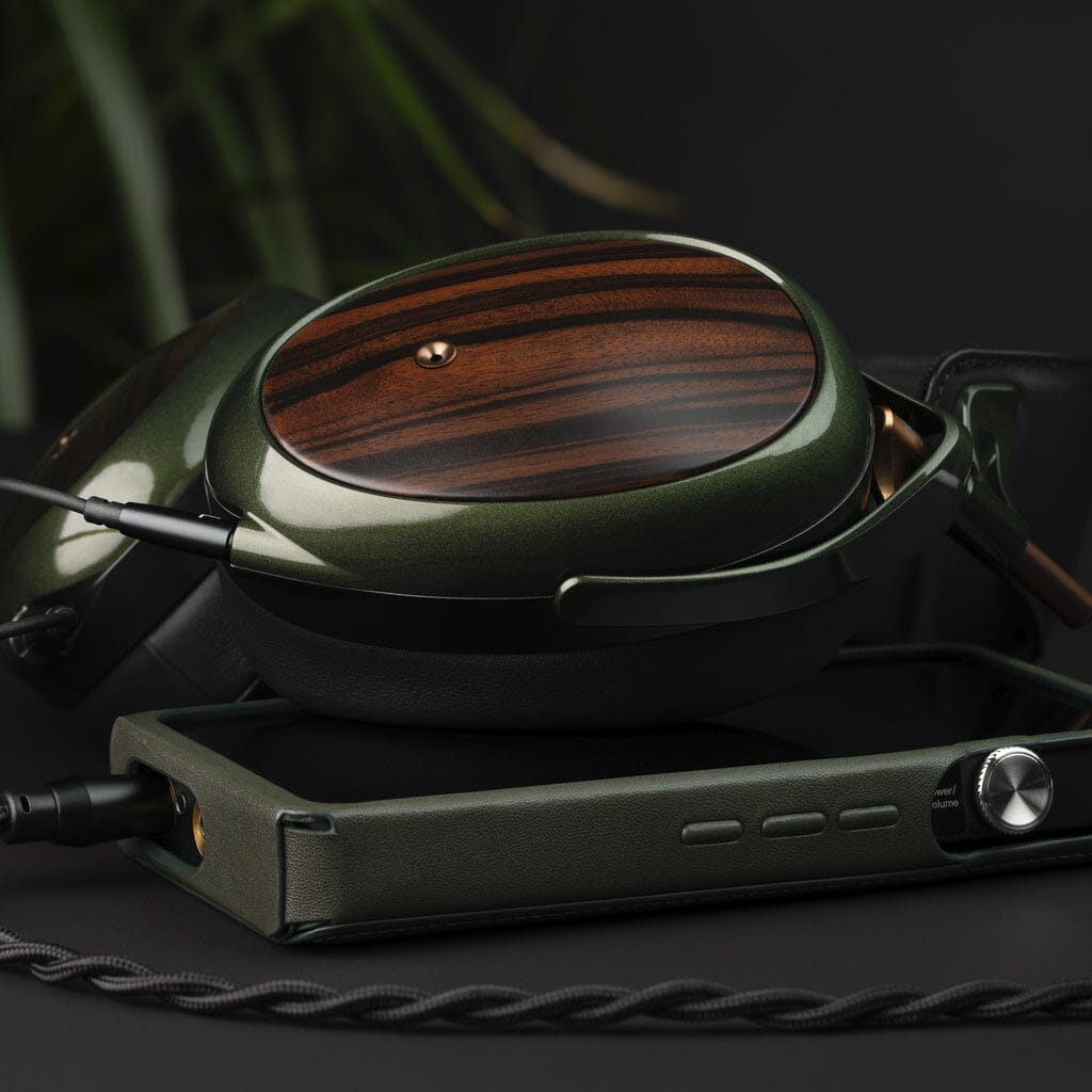 A pair of Meze Audio Strada closed-back headphones with wooden ear cups and a dark green metallic frame rests beside a matching portable audio player on a black surface, with a blurred leafy plant in the background, reflecting refined tonal balance.