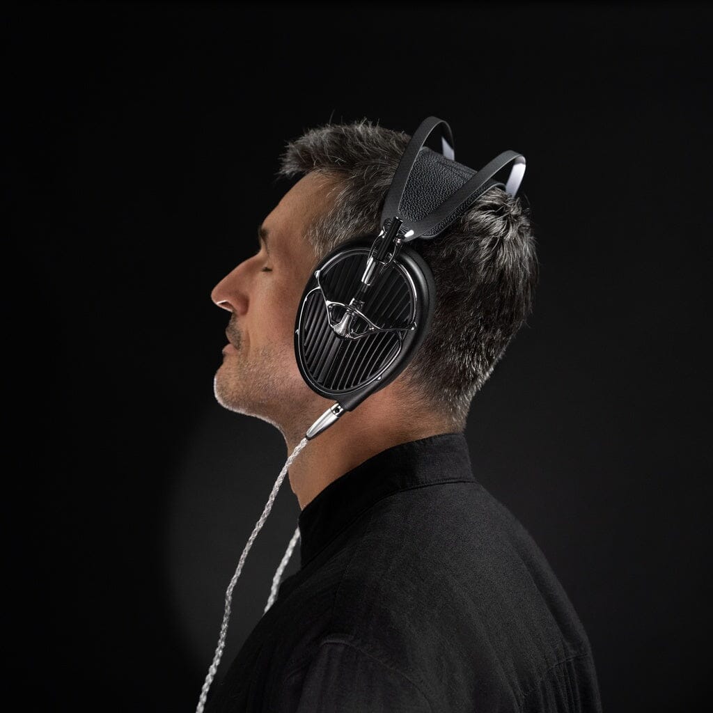 A man with short gray hair, wearing large Meze Audio ARTA Headphones and a black shirt, faces left against a plain black background.
