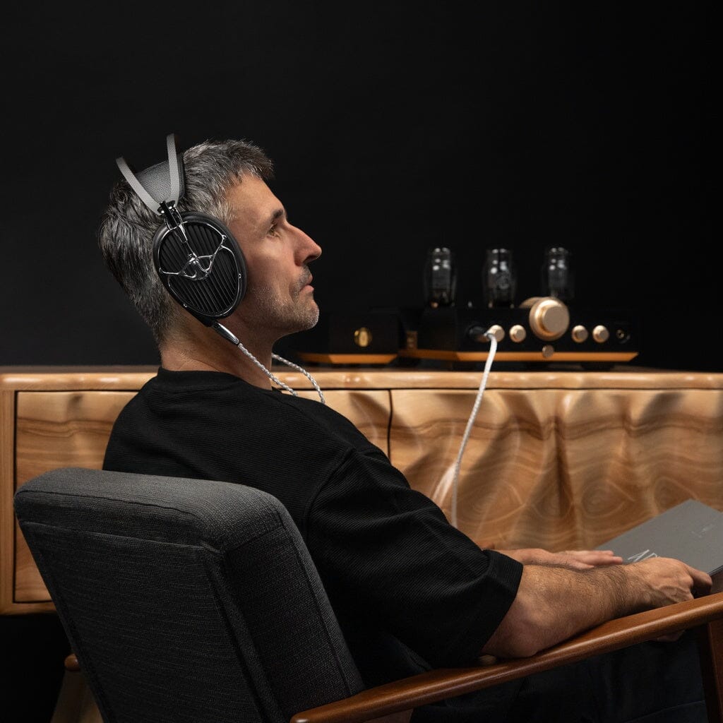 A man sits in a chair wearing Meze Audio ARTA Headphones, looking relaxed. In the background, audio equipment with glowing tubes rests on a wooden cabinet, lending a calm and sophisticated atmosphere.