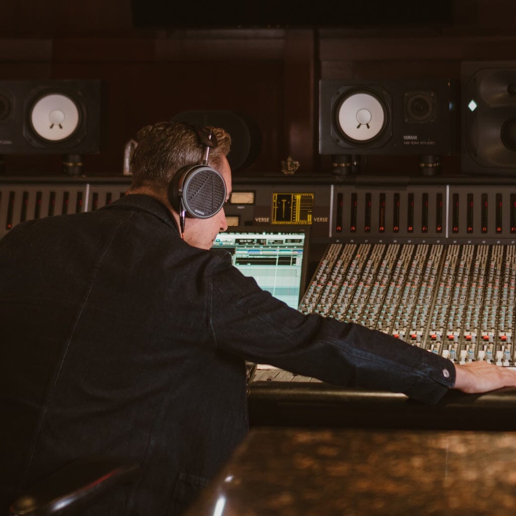 A person wearing Audeze MM-520 Headphones sits at a large audio mixing console in a recording studio, adjusting controls while looking at a computer screen displaying audio tracks.