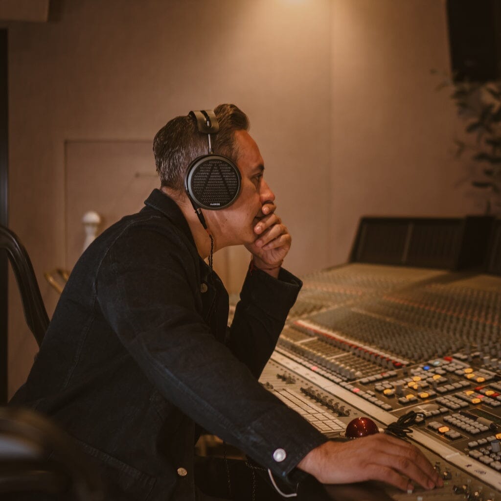 Wearing Audeze MM-520 Headphones, a person sits at a recording studio mixing console, chin resting on their hand as they focus intently on the controls.