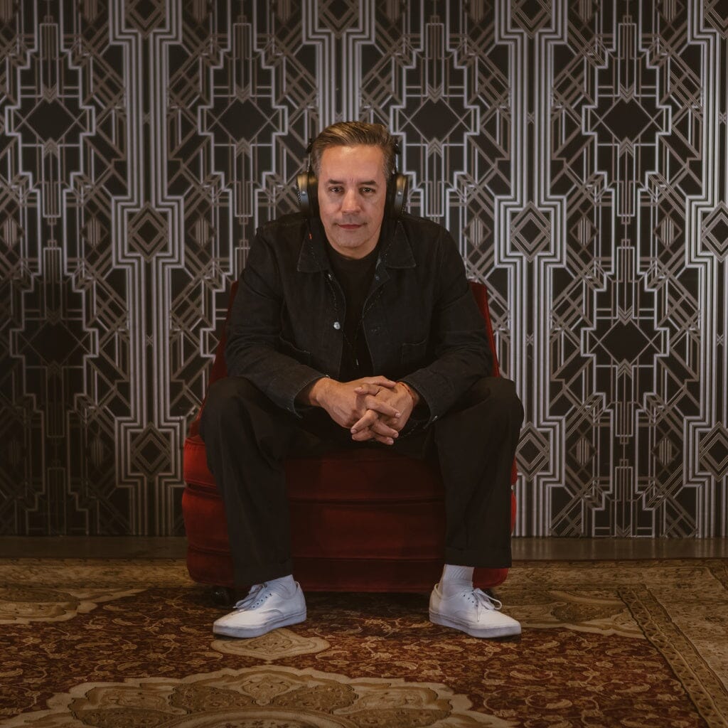 A man in dark clothing, wearing Audeze MM-520 headphones by Audeze, sits on a red chair against a geometric wall and patterned rug, looking directly at the camera.