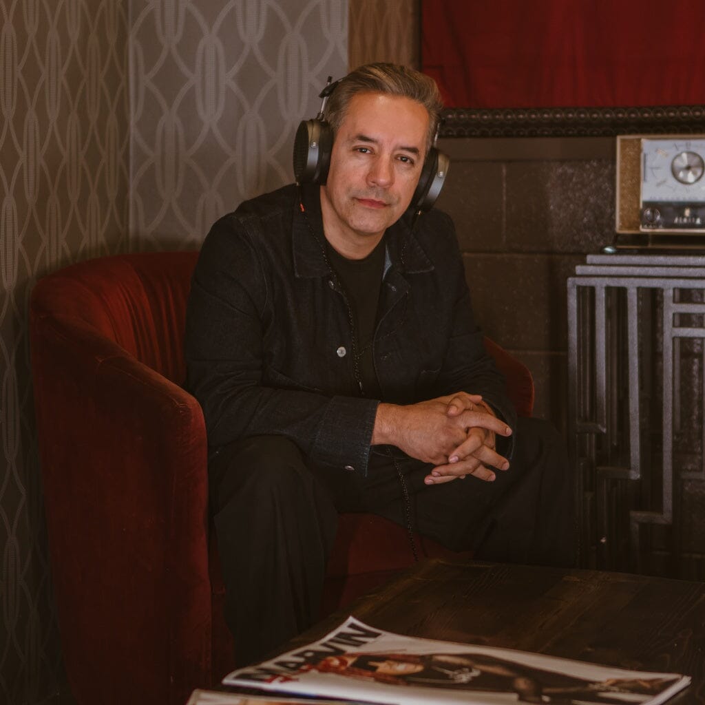 A man with short gray hair, dressed in dark clothes, sits on a red armchair in a dim room with patterned wallpaper, wearing Audeze MM-520 Headphones by Audeze. A magazine and vintage radio rest on a table nearby.