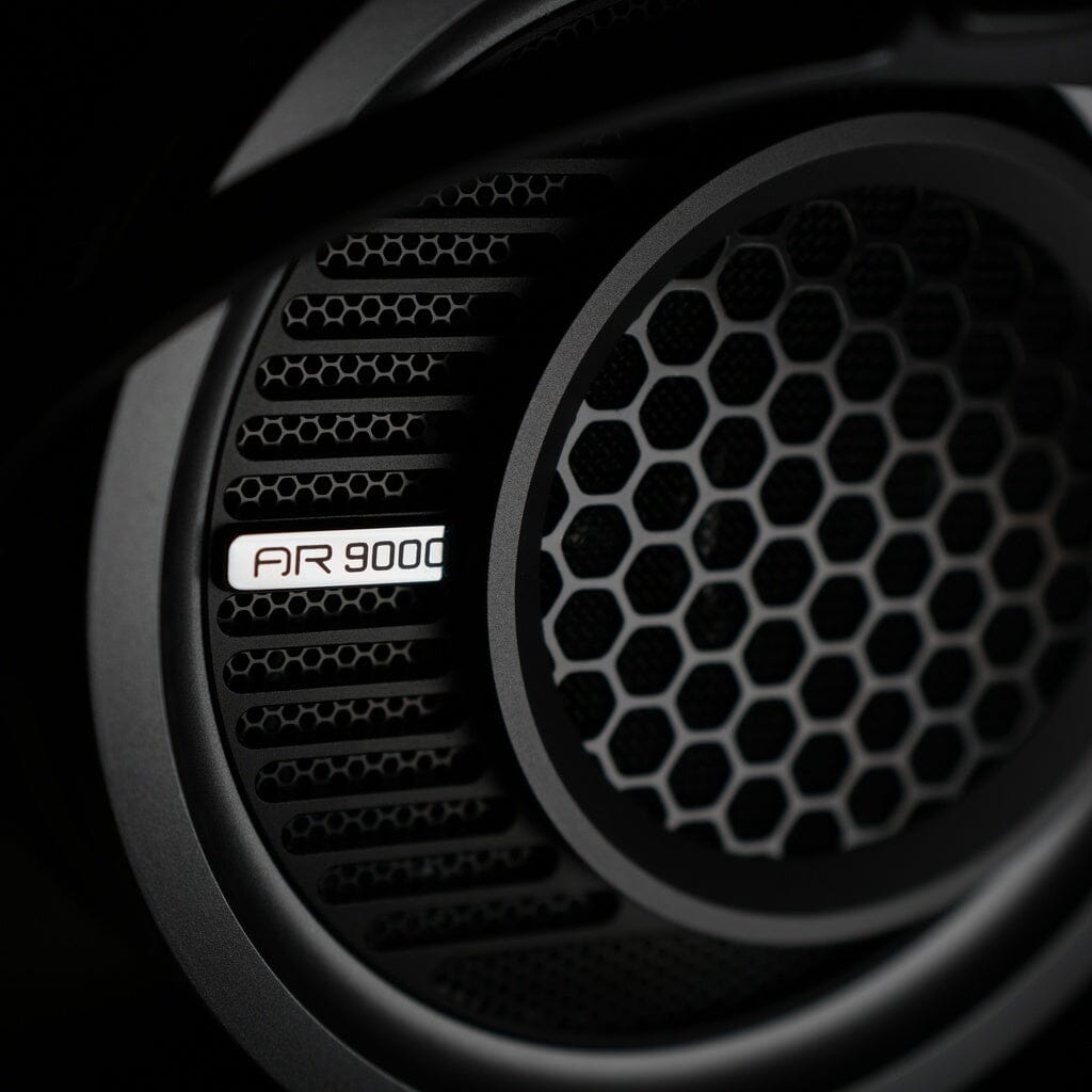 Close-up of a headphone speaker with a black honeycomb grille pattern and the Aune AR9000 label visible on the side. The image is sharply focused on the details of the grille and label, emphasizing the custom 50mm dynamic driver engineered by Aune Audio for low distortion—demonstrating the precision characteristic of Aune AR9000 Headphones.
