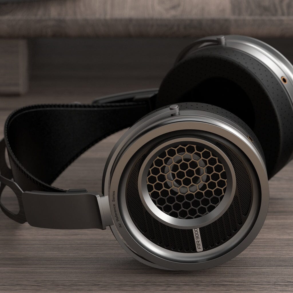 A pair of Aune AR9000 headphones from Aune Audio, featuring custom 50mm dynamic drivers and a distinctive metal mesh grille on the ear cups, rests on a wooden surface with a shelf visible in the background. Engineered for low distortion, these high-end over-ear headphones deliver exceptional audio clarity and detail.