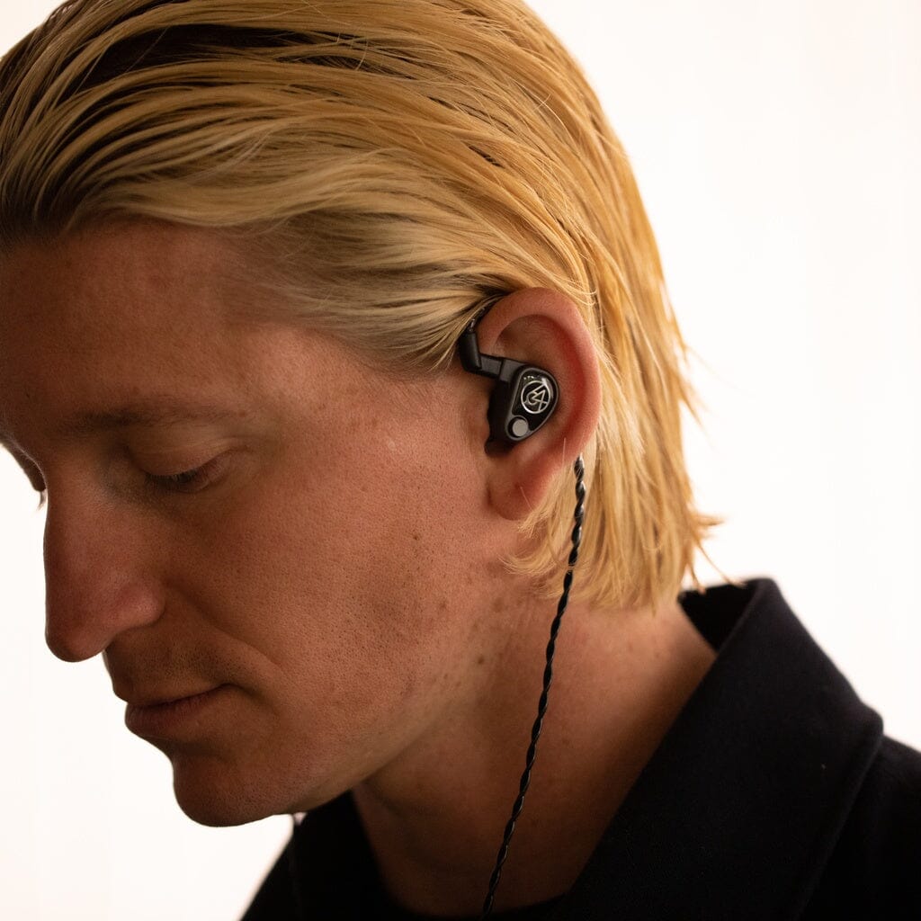 A blond-haired person wears black 64 Audio U6t In-Ear Headphones with a visible logo, looking down with a neutral expression against a plain, light background.