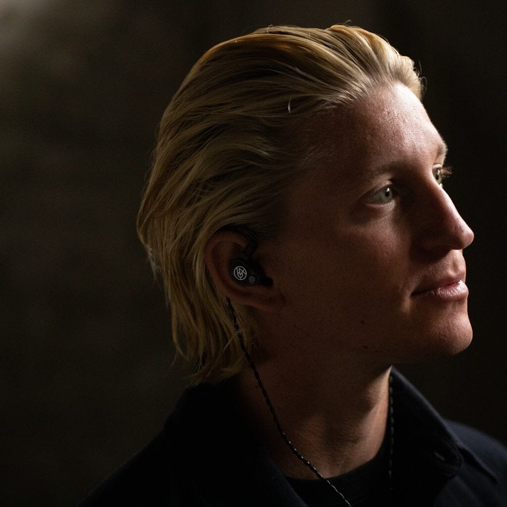 A person with light skin and blonde hair looks upward while wearing wired black 64 Audio U6t In-Ear Headphones. The dark, blurred background highlights their face and showcases the high-quality 64 Audio audiophile monitors.