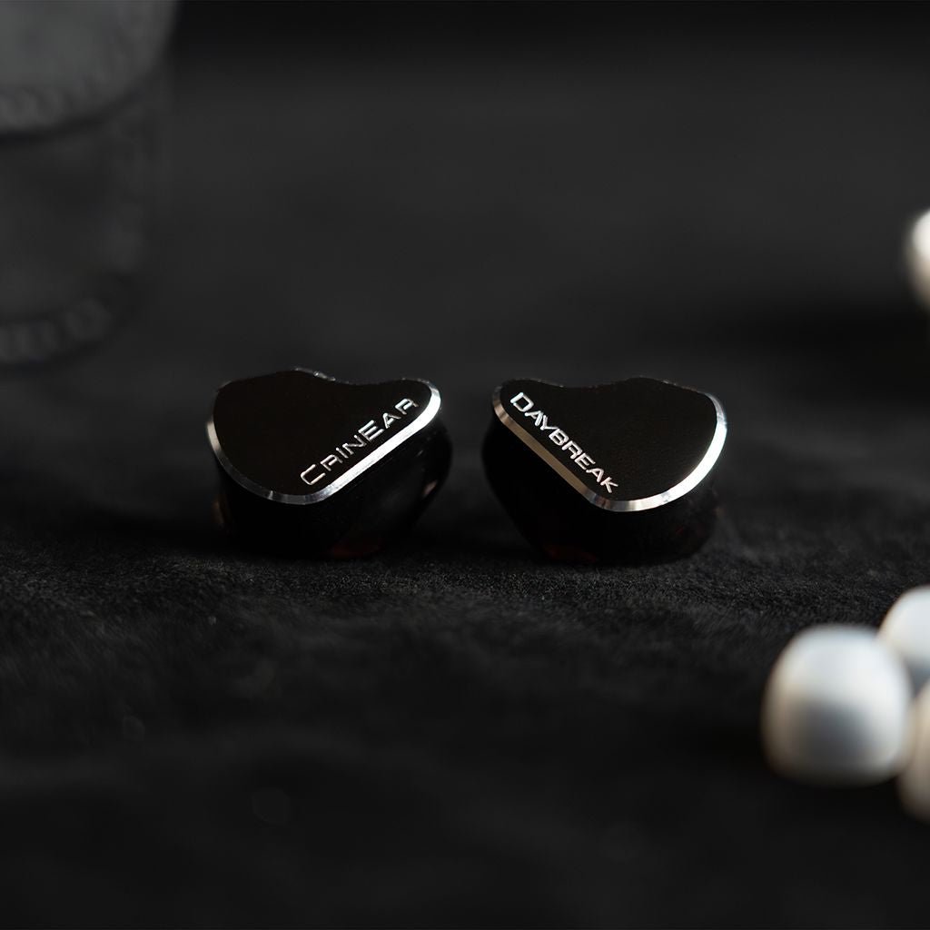 A pair of Crinear Daybreak In-Ear Headphones by Crinear with white ear tips rest on a dark surface, offering audiophile-grade sound as highlighted on headphones.com.