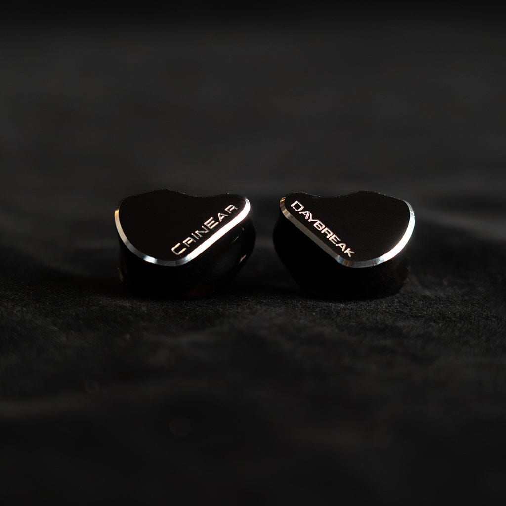 Two black Crinear Daybreak In-Ear Headphones, labeled “CRIN EAR” and “DAYBREAK,” rest on a dark surface with reflective metal trim—crafted for audiophile sound and available at headphones.com. Brand: Crinear.