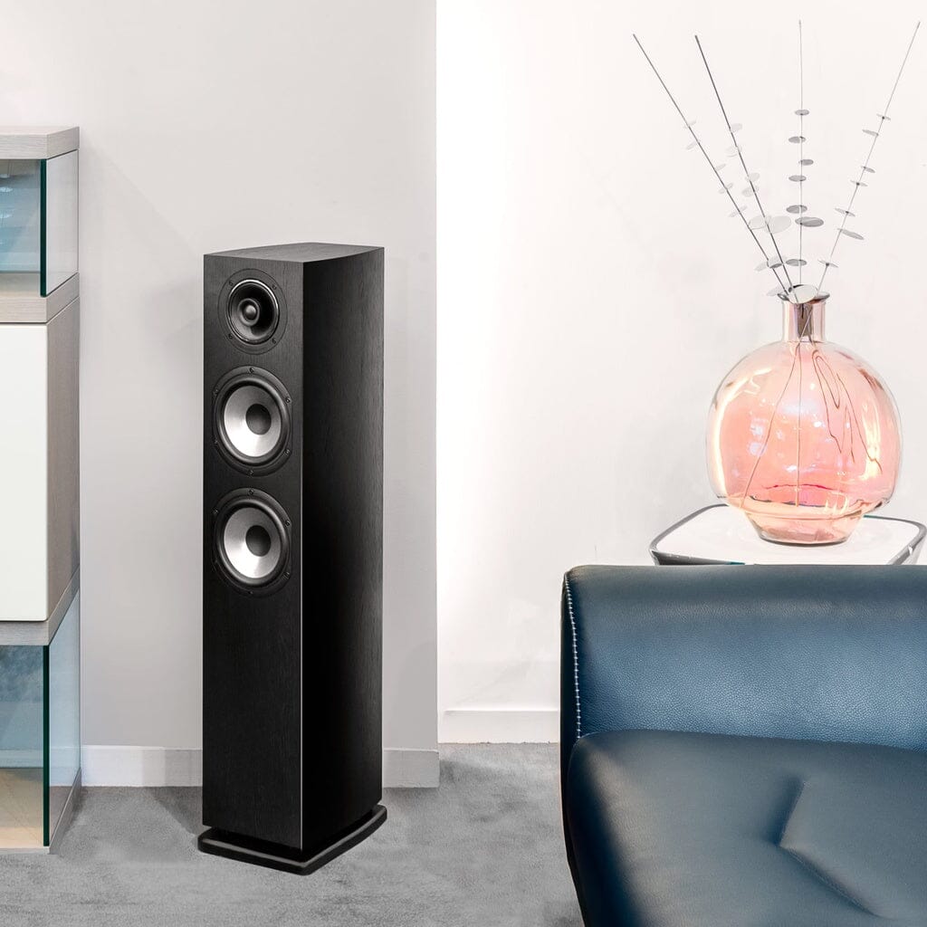 A modern living room with a tall black Cabasse JERSEY MC170 floorstanding speaker, a glass vase with decorative sticks on a side table, and a blue leather sofa on grey carpeted flooring.
