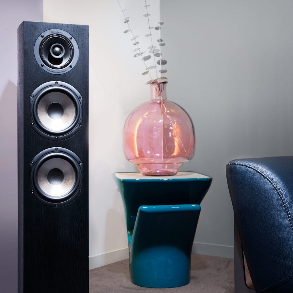 A sleek black Cabasse JERSEY MC170 Floorstanding Speaker stands beside a blue chair and a table with a large pink glass vase in a modern, minimalist room.