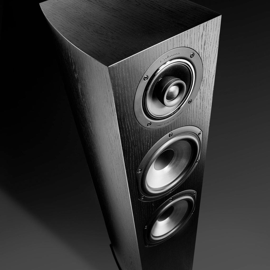 A close-up angled view of the Cabasse JERSEY MC170 Floorstanding Speaker by Cabasse shows its tall wood-grain design, three circular drivers in a vertical row, and high-powered woofers against a dark background.