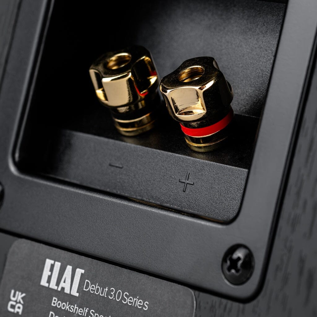 Close-up of two gold speaker terminals, one with a red ring, showing polarity symbols on the back of an ELAC Debut 3.0 DB63 6.5" Bookshelf Speaker featuring an aramid fiber woofer and aluminum dome tweeter.