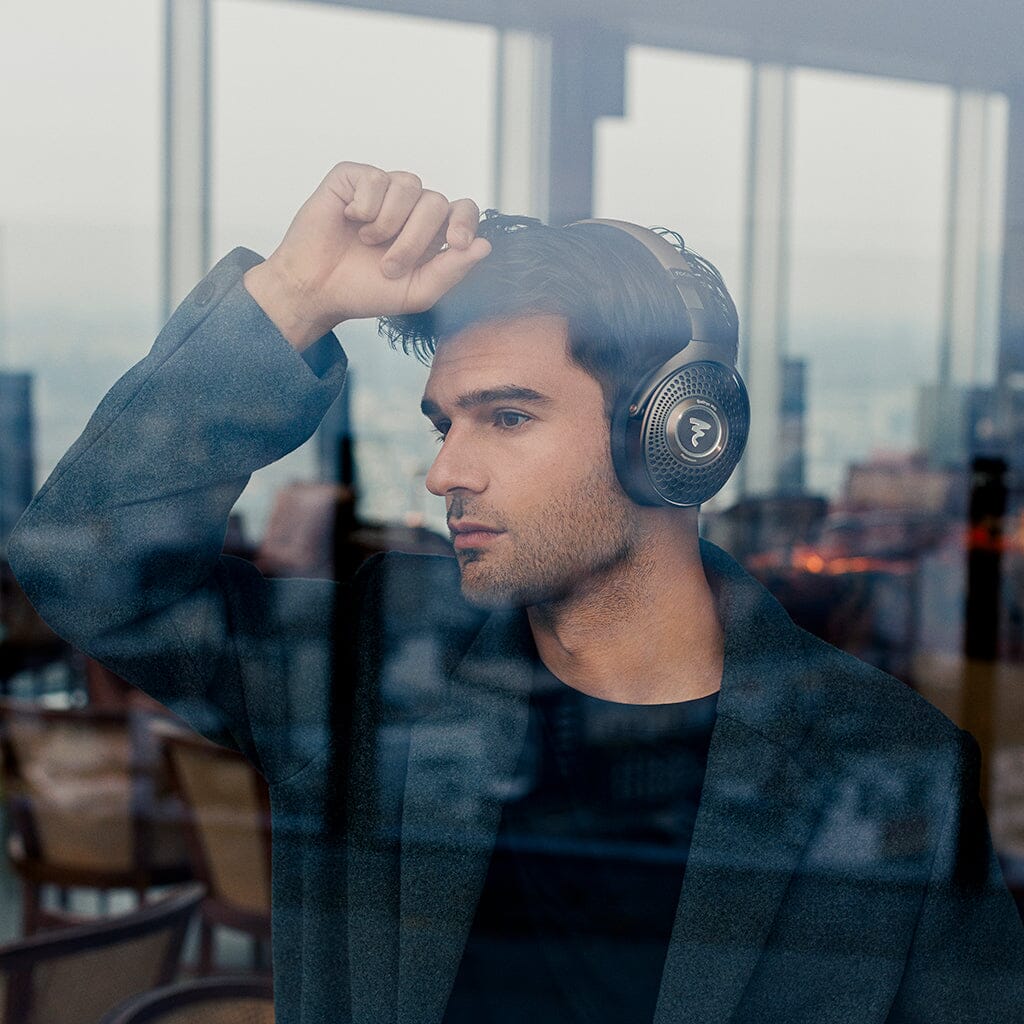 A man in a dark coat, wearing Focal Bathys MG Wireless Headphones by Focal, gazes thoughtfully out a window with his hand on his forehead. Outside, the scene is blurred and reflections shimmer on the glass.