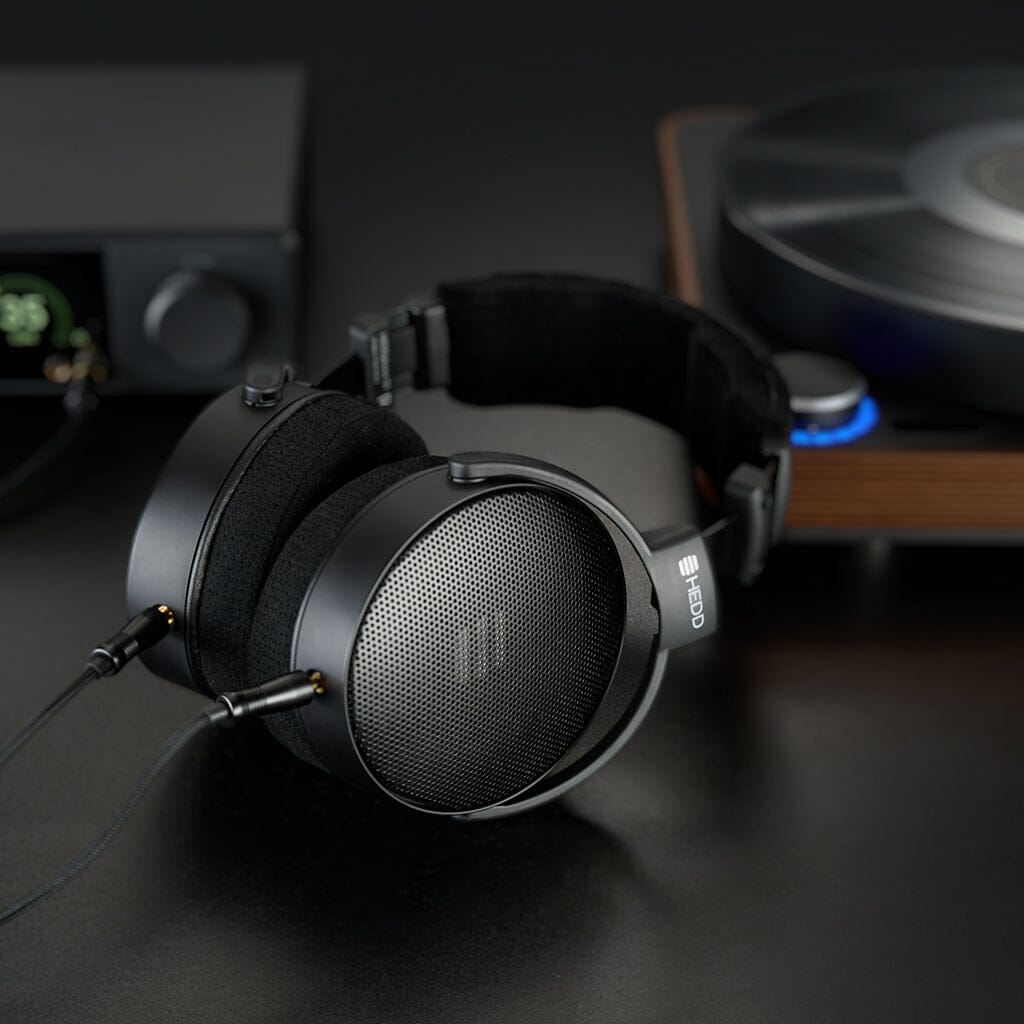 A pair of HEDD Audio HEDDphone D1 headphones with mesh ear cups and a Thin-Ply Carbon Diaphragm rest on a dark surface near a wooden turntable and audio amplifier, connected to the equipment.